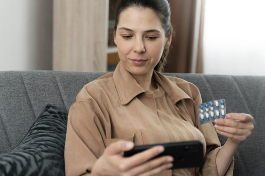 A woman sitting on a couch, holding a blister pack of UTI tablets in one hand and using a smartphone in the other, suggesting engagement with women's urinary health.