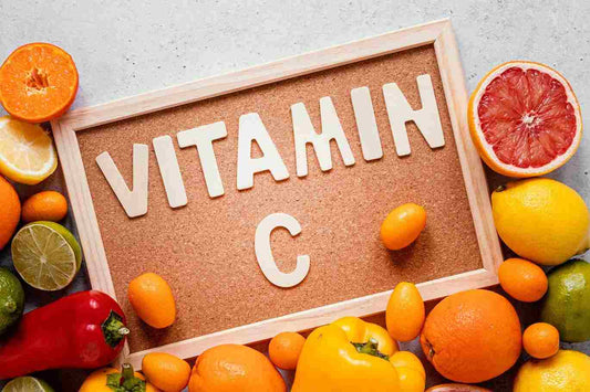 A corkboard with the text "VITAMIN C" spelled out in white letters, surrounded by various citrus fruits, including oranges, lemons, and grapefruits, along with a red bell pepper and yellow-orange fruits, arranged on a light gray background.