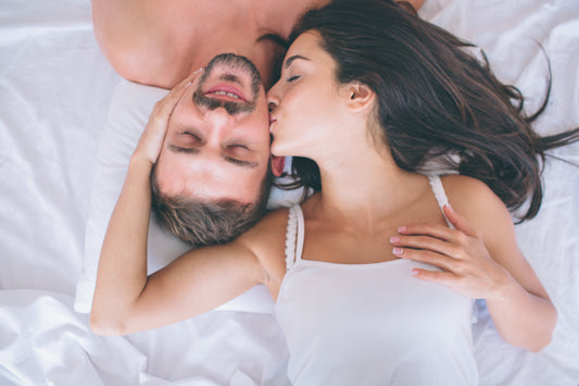 An image of a woman giving a gentle kiss to a man lying on a bed, with both of them relaxed and dressed in casual white clothing.