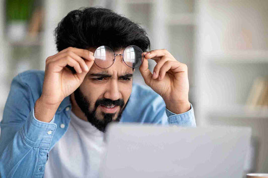 A man adjusting his glasses while looking at a laptop screen with a focused expression. The setting appears to be a well-lit room with bookshelves in the background, emphasizing the theme of visual engagement.