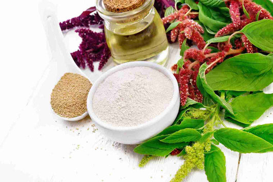 A white bowl filled with powdered collagen supplement sits on a wooden surface, alongside a spoon of brown powder and fresh green herbs. A glass jar of oil is in the background, with colorful herbs arranged around.