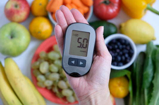 An image of a hand holding a glucose meter showing a reading of 5.0, surrounded by various fresh fruits and vegetables.