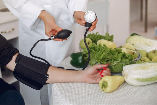 A healthcare professional measuring blood pressure on a patient's arm using a sphygmomanometer, with a variety of fresh vegetables displayed on a table in the background.