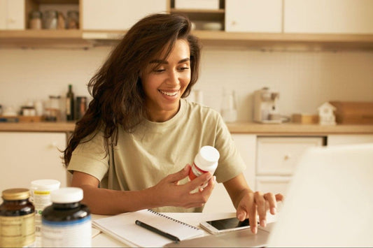 A woman sitting at a desk in a kitchen, smiling while holding a bottle of Daily Nutrition Supplements and looking at her laptop. Various supplement bottles are visible on the table.