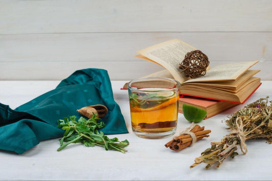A glass of herbal tea with herbs and spices, including cinnamon sticks, placed next to an open book on a white surface. A green fabric and additional dried herbs are visible, suggesting a focus on herbal supplements.