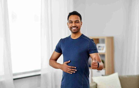 A smiling man in a blue t-shirt stands in a bright room, giving a thumbs-up while placing his hand on his stomach. 