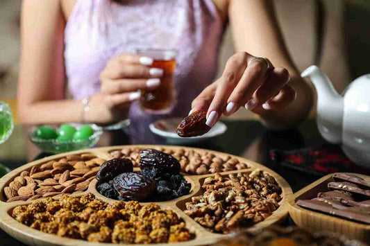 A woman holding a date while reaching for a platter of mixed nuts and sweets, with a glass of beverage in her other hand.